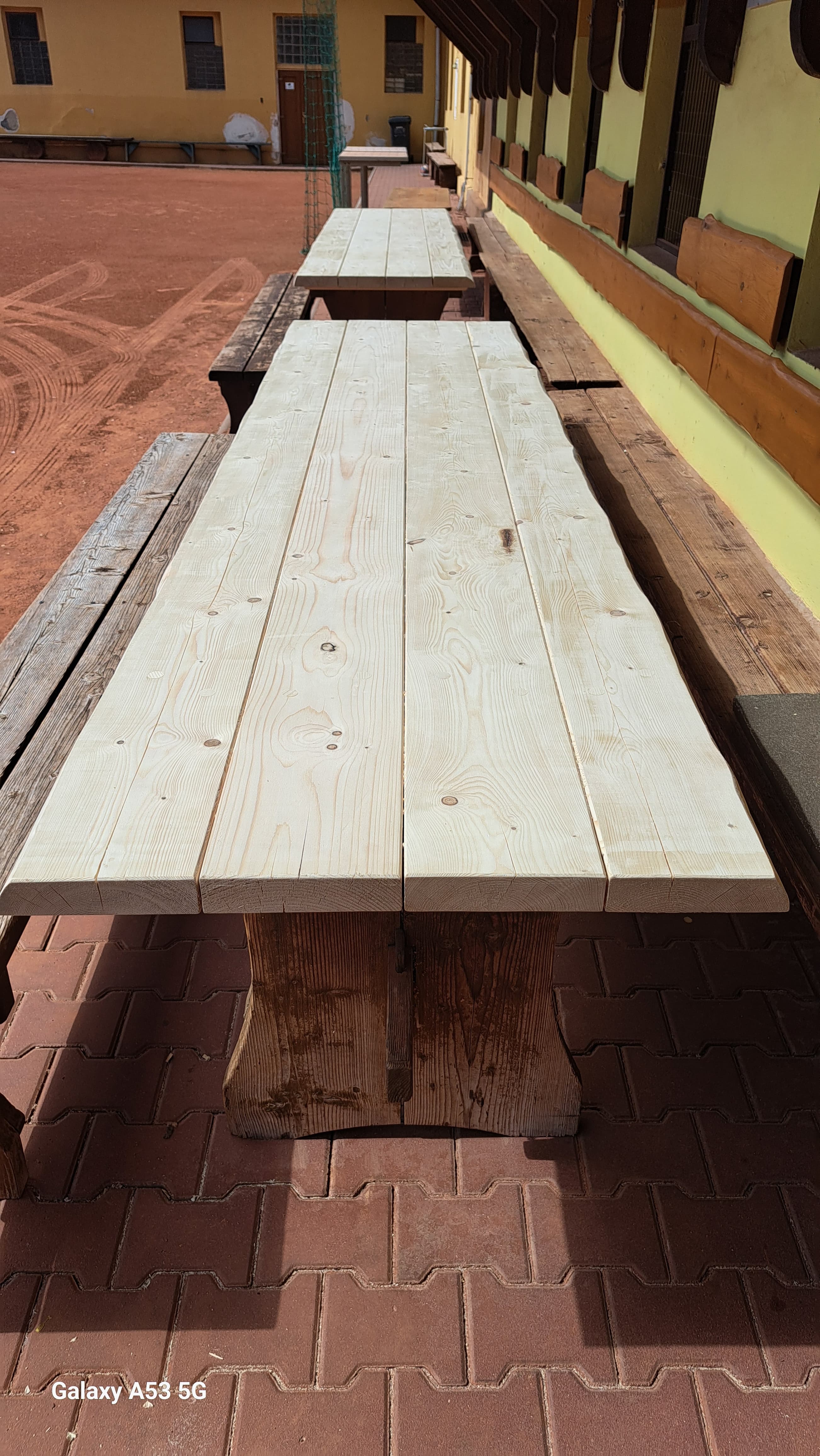 Long unfinished light-colored wooden table and benches on a red brick patio outdoors.