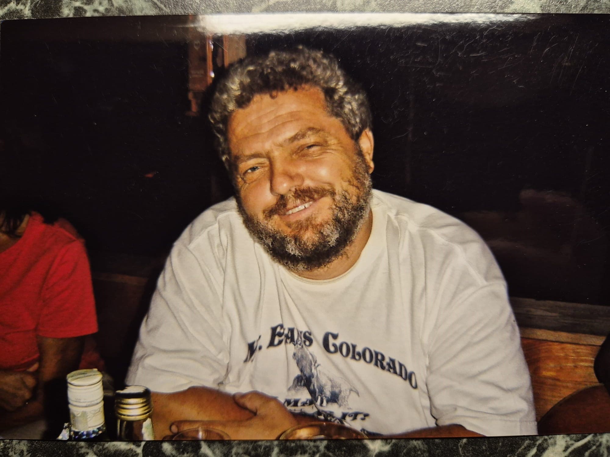Bearded man smiling while wearing a white Mt. Evans Colorado t-shirt at a restaurant.