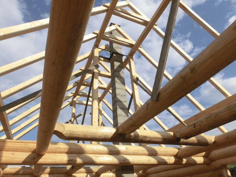 Wooden roof rafters and log beams frame a concrete chimney against a cloudy blue sky.